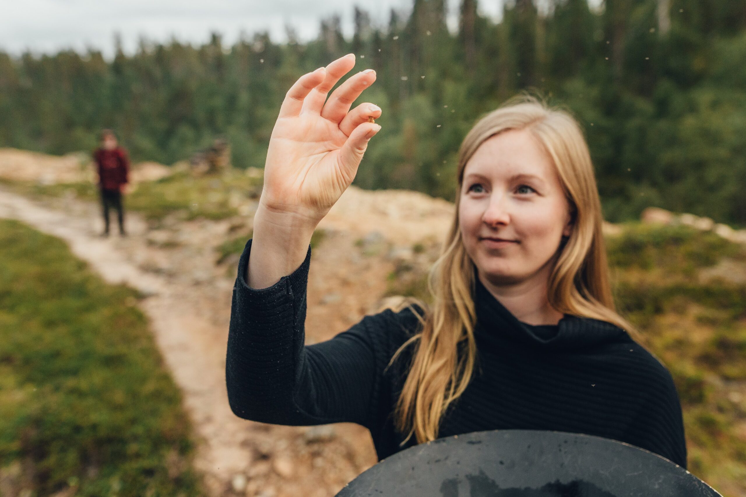 Pitkät vaaleat hiukset omaava nainen pitää pientä esinettä sormiensa välissä ja tutkii sitä tarkasti ulkona metsäisessä maisemassa, jossa toinen henkilö seisoo polulla hänen takanaan.