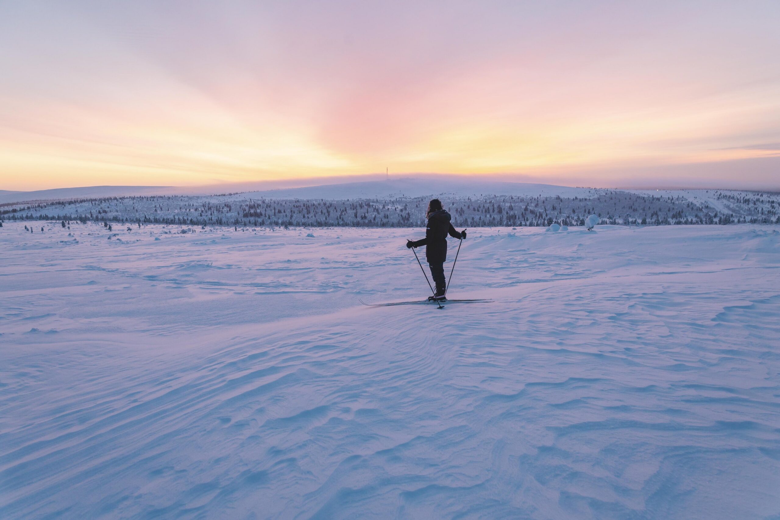 Henkilö hiihtää yksin lumipeitteisessä maisemassa auringonlaskun aikaan, kun taivaan pehmeät vaaleanpunaiset ja oranssit sävyt täyttävät taivaan ja valaisevat hellästi talvisen maiseman.