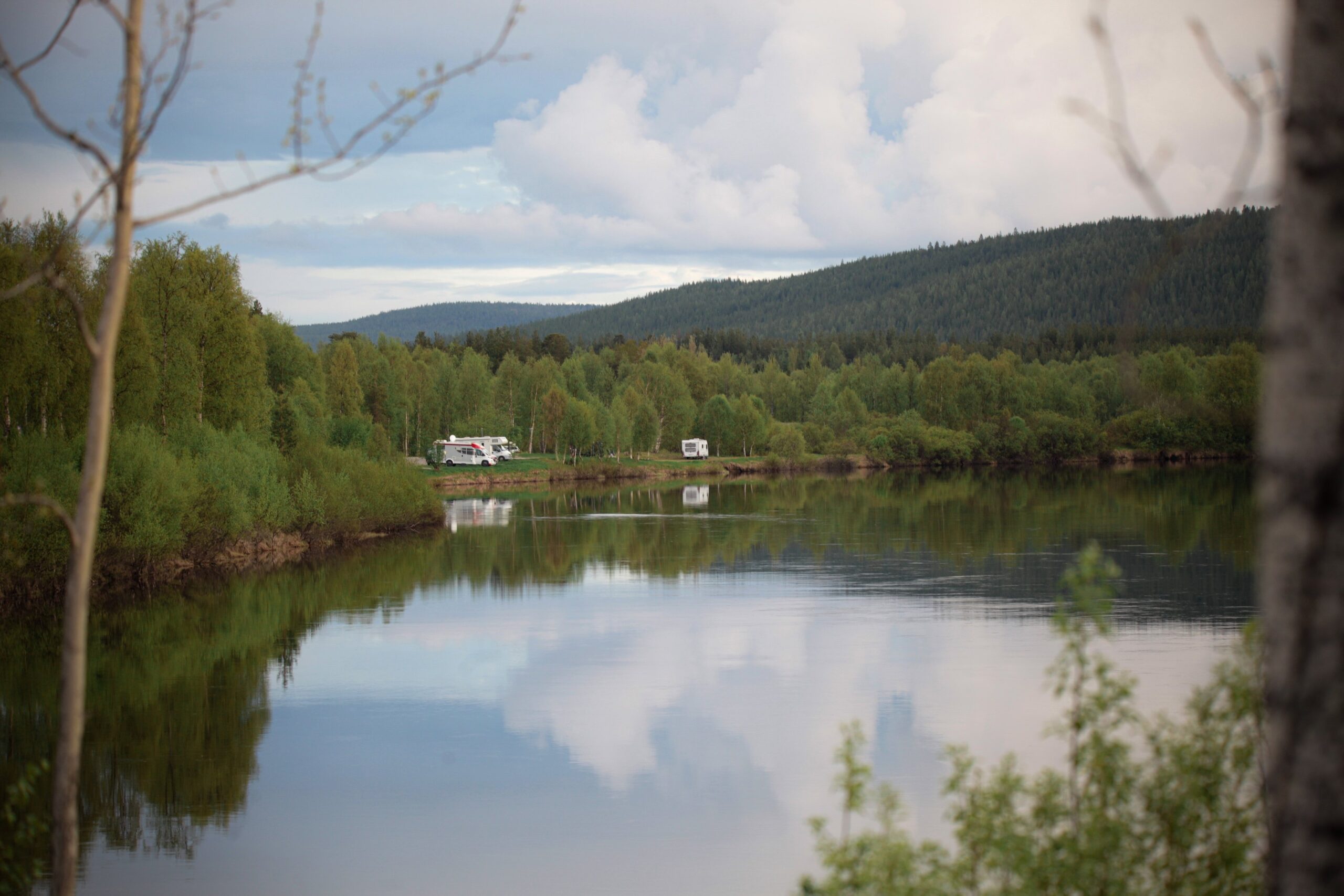 Kaksi asuntoautoa on pysäköity rauhallisen, heijastavan järven rannalle, jota ympäröivät tiheät vihreät puut ja metsäiset kukkulat pilvisen taivaan alla. Maisema on rauhallinen ja luonnollinen, ja vesi heijastaa maisemaa.
