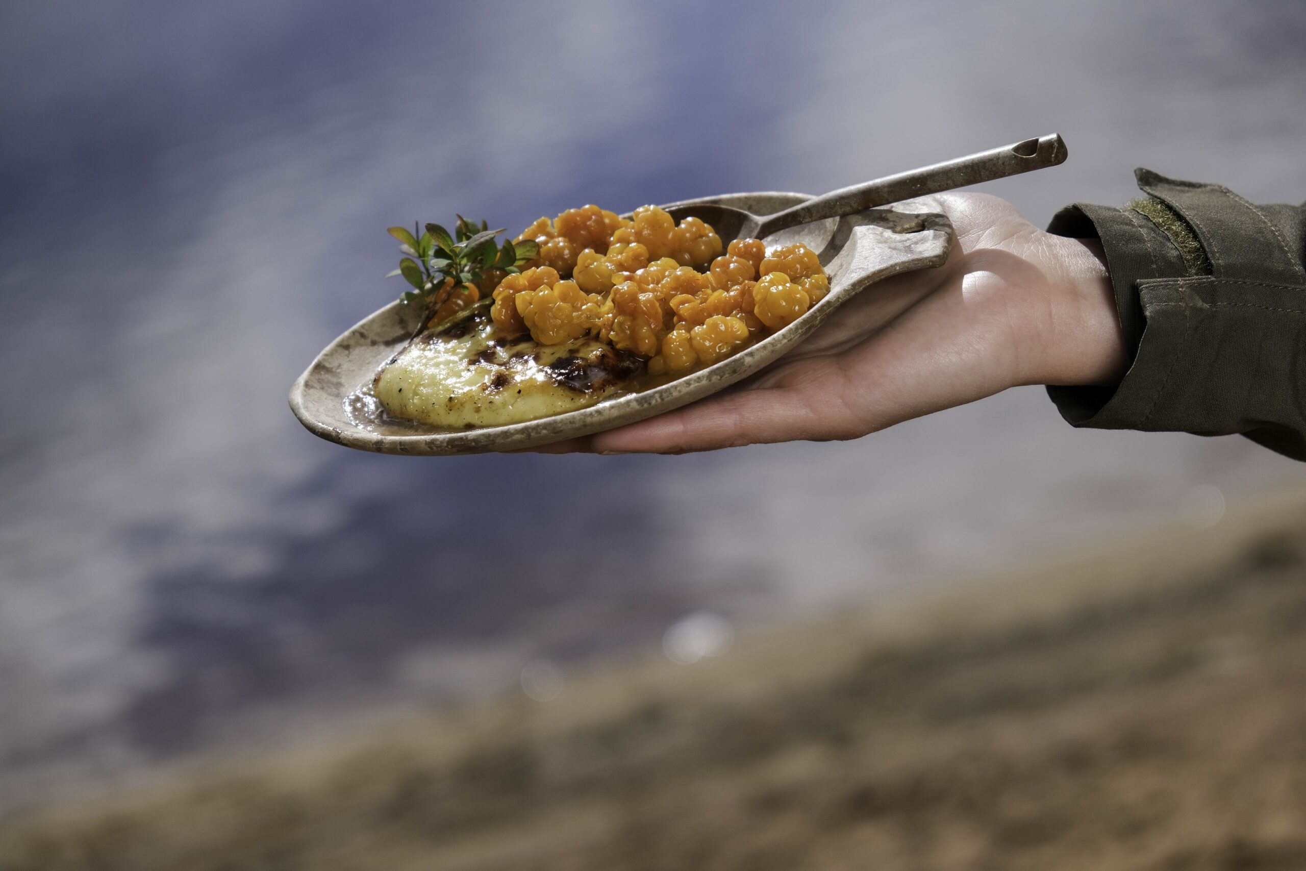 Kädessä on maalaismainen lautanen ja lusikka, jonka päällä on kirkkaan oransseilla pilvenmarjoilla koristeltu lautanen, joka on koristeltu vihreillä lehdillä, ja taustalla on sumea ulkoilmakuva.