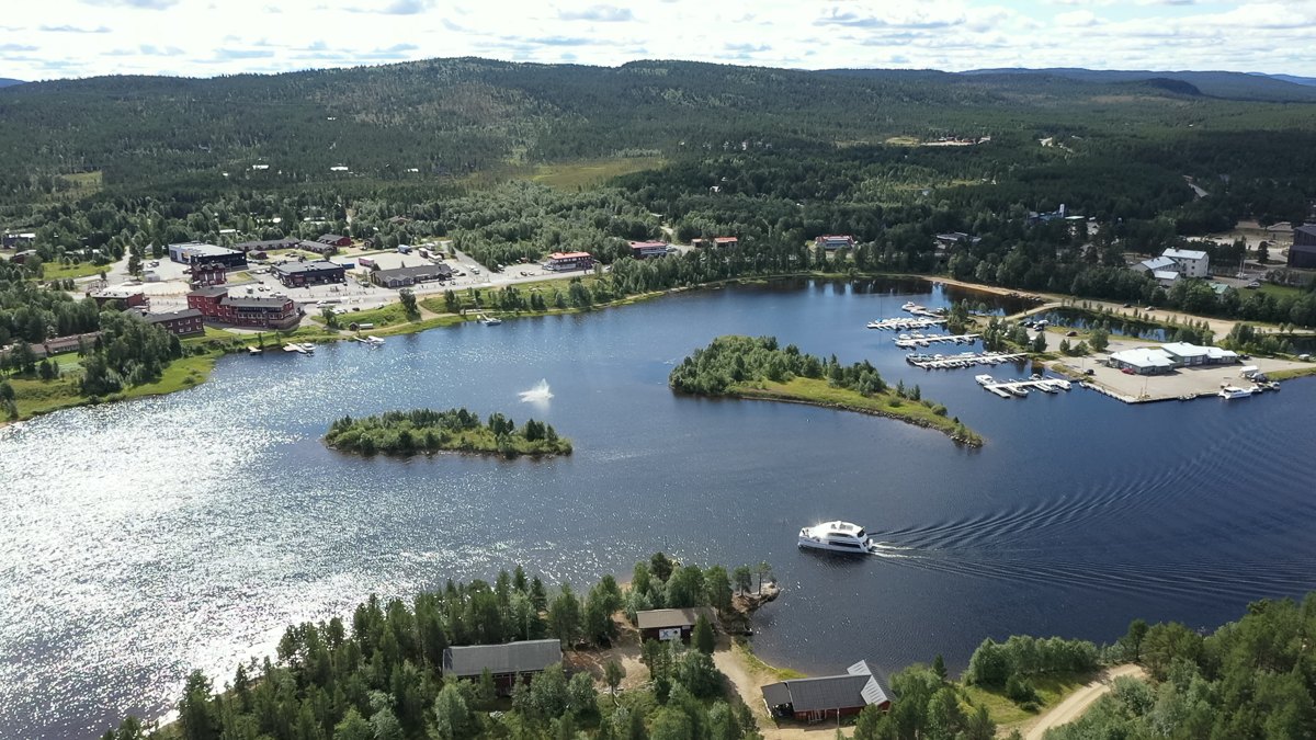 Ilmakuva rauhallisesta järvestä, jossa on pieniä saaria, vesillä risteilevä vene ja venesatama, jossa on telakoituja veneitä. Järven ympärillä on vihreitä metsiä, hajallaan olevia rakennuksia ja pieni kaupunki.