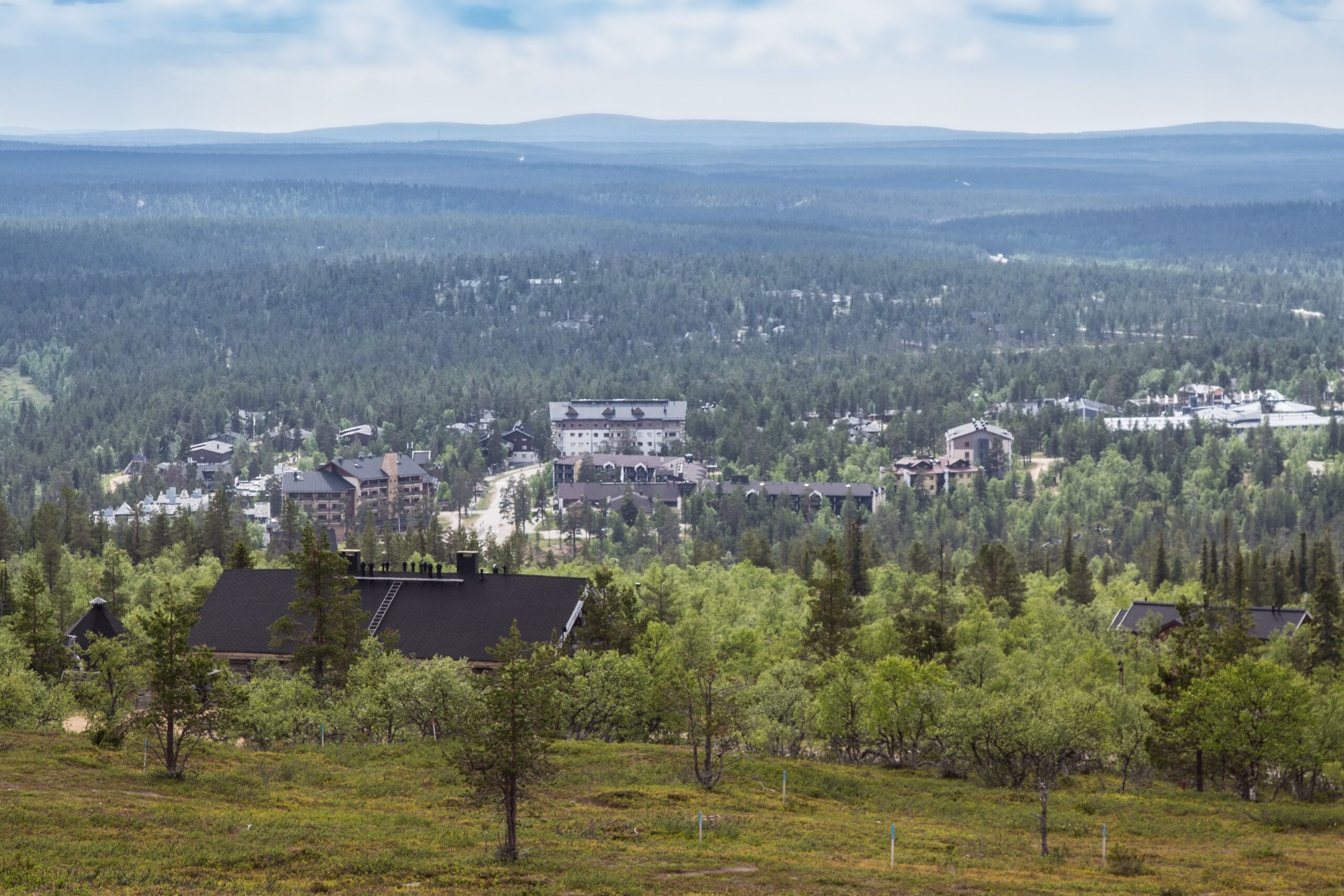 Ein Panoramablick auf eine bewaldete Landschaft mit mehreren Gebäuden zwischen den Bäumen, sanften Hügeln im Hintergrund und einem bewölkten blauen Himmel darüber.