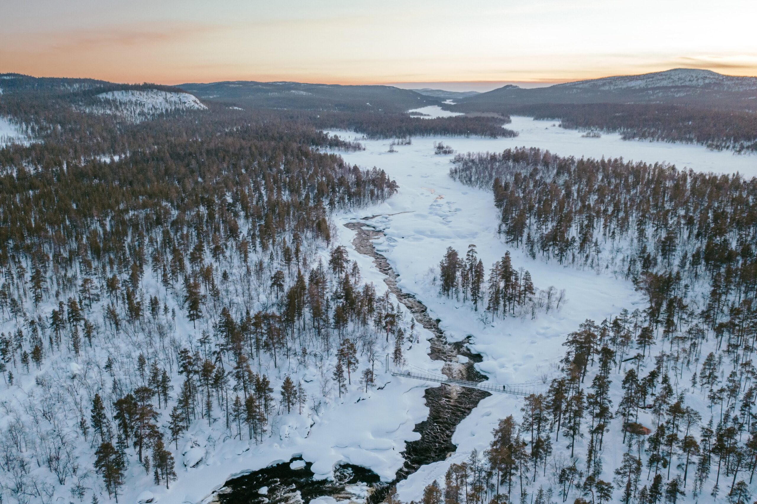 Luftaufnahme einer verschneiten Waldlandschaft mit einem sich durch die Bäume schlängelnden Fluss, der teilweise zugefroren ist, unter einem blassen Himmel bei Sonnenuntergang oder Sonnenaufgang. Schnee bedeckt den Boden und die Hügel in der Ferne.