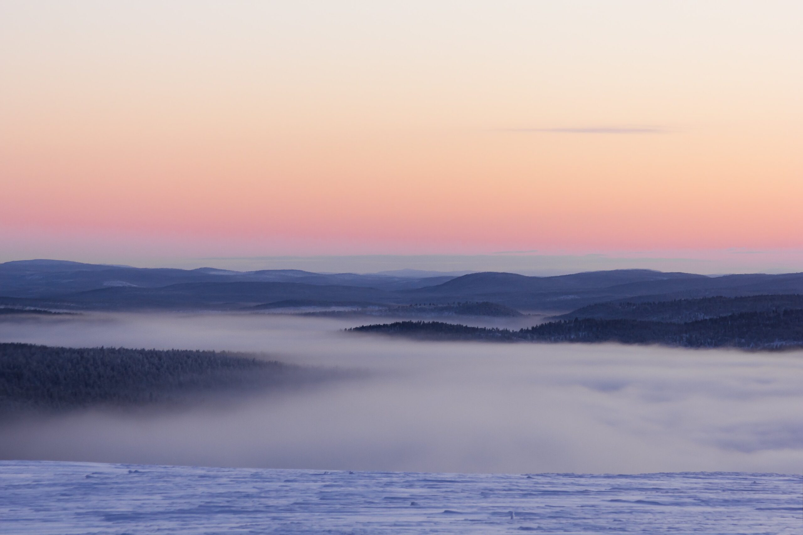 Lumipeitteinen maisema, jossa puiden peittämät kukkulat ovat osittain matalan sumun peitossa. Taivas on sävytetty vaaleanpunaiseksi ja oranssiksi, mikä viittaa auringonnousuun tai -laskuun, ja pehmeät pastellivärit hallitsevat seesteistä talven maisemaa.