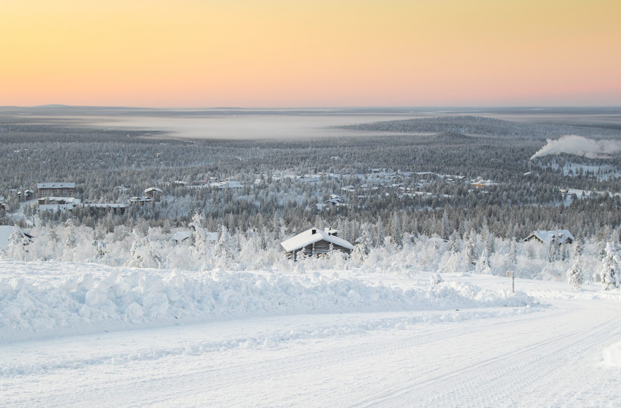 Luminen maisema, jossa talot ovat lumipeitteisten puiden keskellä pastellinvärisen oranssin ja keltaisen taivaan alla auringonnousun tai auringonlaskun aikaan, ja taustalla on kaukaisia kukkuloita ja sumukerros.