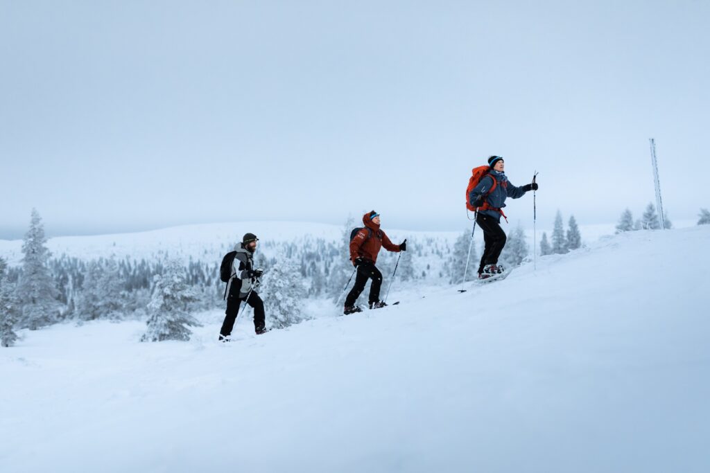 Kolme talvivaatteisiin pukeutunutta ihmistä vaeltaa syvässä lumessa hiihtosauvojen kanssa ylämäkeen lumisella, puiden reunustamalla vuorenrinteellä pilvisen, pilvisen taivaan alla.