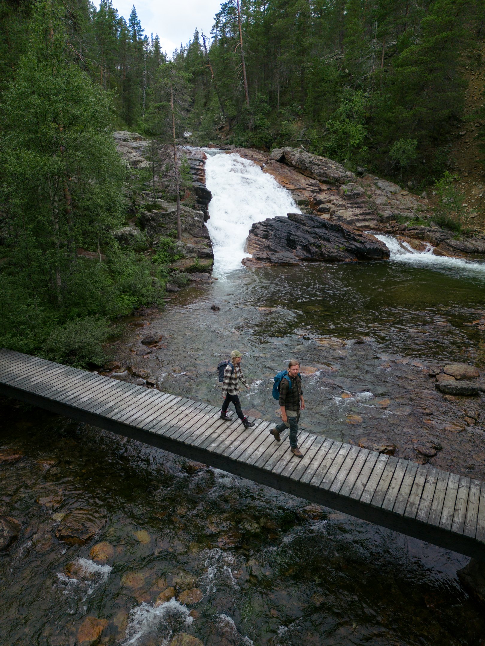 Two people walk across a wooden bridge over a river, with a waterfall and dense green forest in the background. The scene appears serene and natural, suggesting a hiking or outdoor adventure.