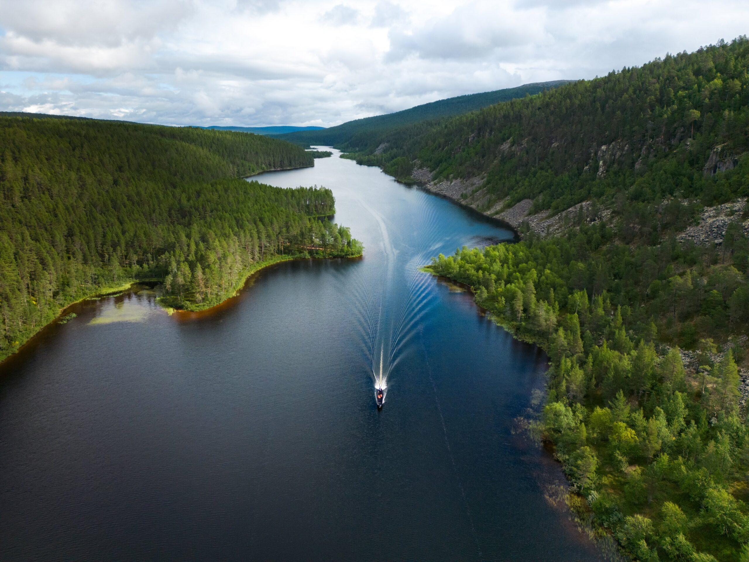 Pieni vene aiheuttaa aaltoja liikkuessaan kapealla joella, jota ympäröi tiheä, vihreä metsä ja kukkulat pilvisen taivaan alla.