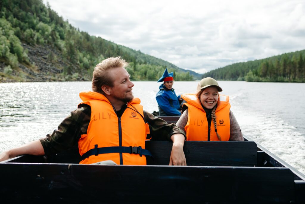 Kolme oransseihin pelastusliiveihin pukeutunutta ihmistä ajaa veneellä rauhallisella joella, jota ympäröivät metsäiset kukkulat, hymyillen ja nauttien ulkoilmasta pilvisen taivaan alla.
