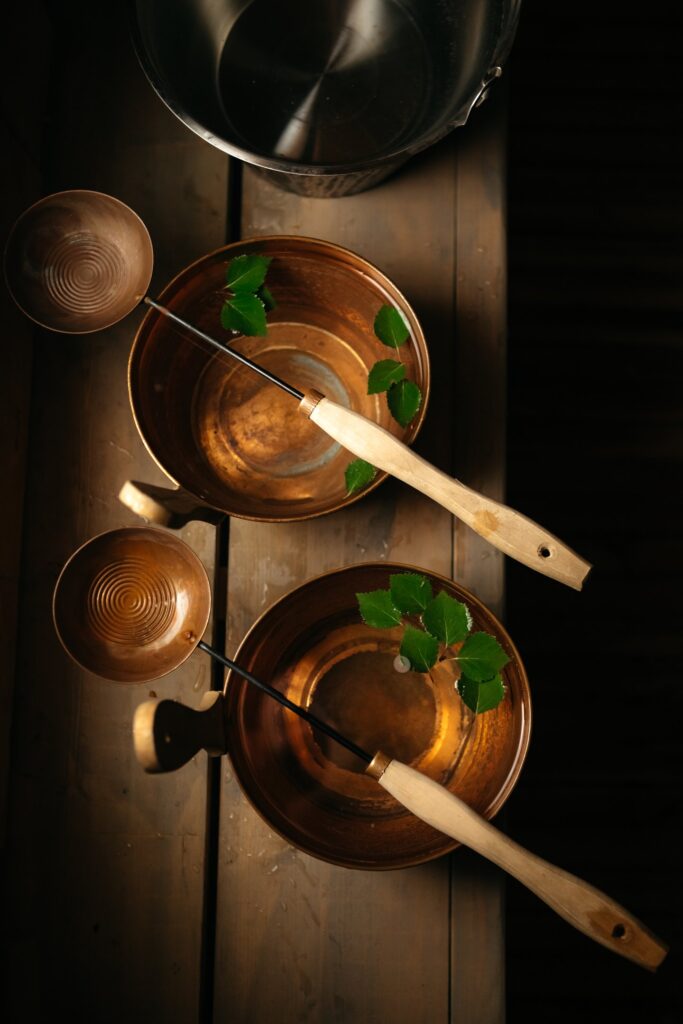 Two wooden ladles rest on copper bowls filled with water and green leaves, placed on a wooden surface, with a metal bucket in the background, all lit by warm, natural light.