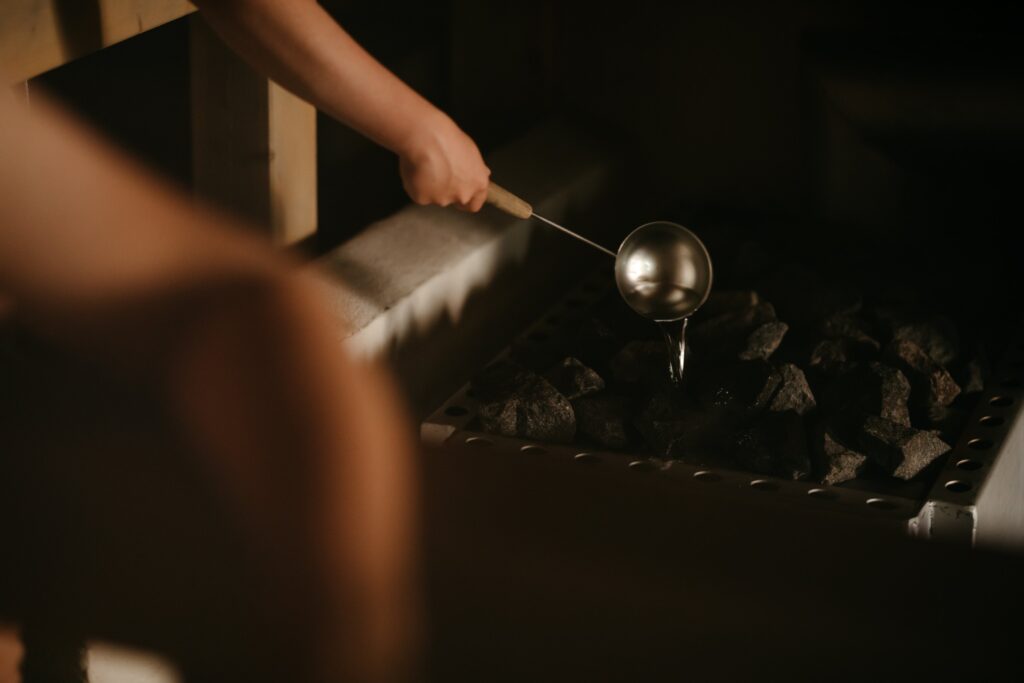 A person’s hand pours water from a ladle onto hot sauna stones, creating steam in a dimly lit wooden sauna room.