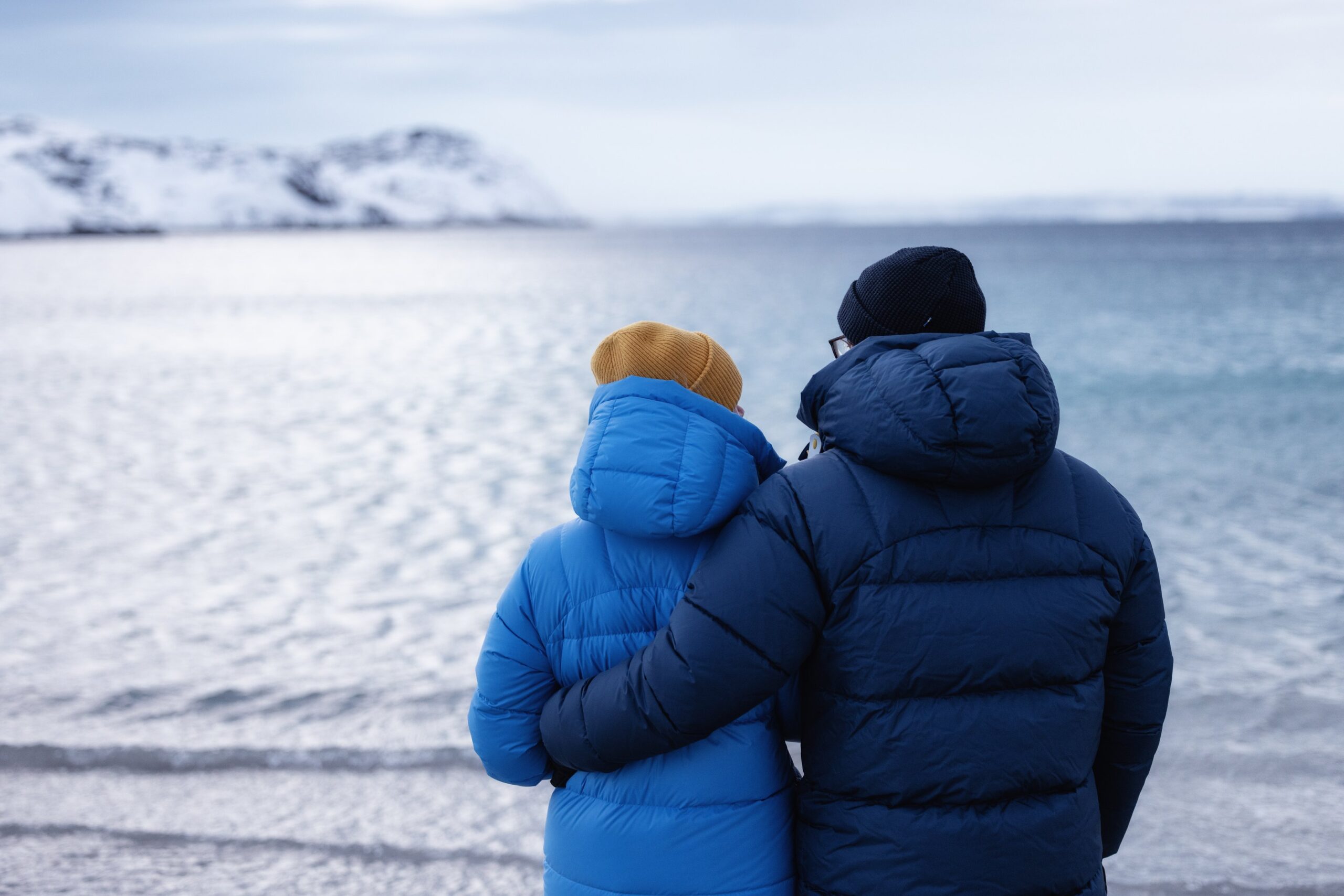 Two people dressed in winter jackets and hats stand on a snowy shore, facing a calm, icy body of water with snow-covered hills in the background. One person has their arm around the other.