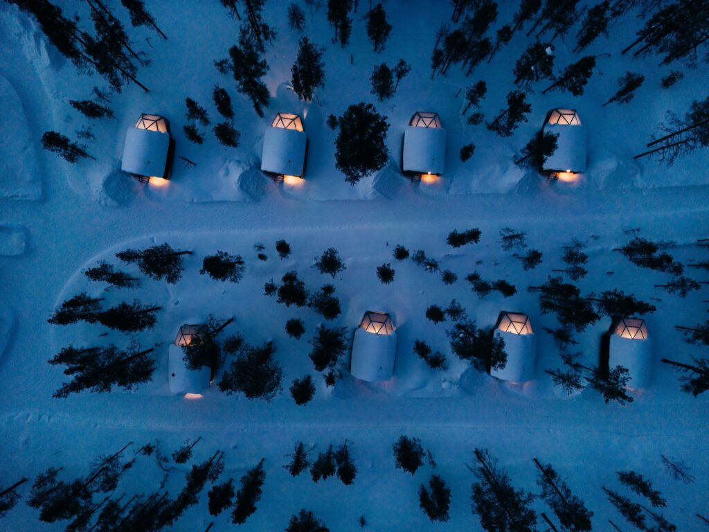 Ilmakuva useista lasikattoisista mökeistä, joiden sisällä palaa lämpimät valot ja joita ympäröi lumen peittämä maa ja puut hämärässä tai yöllä. Talvimaisemassa mutkittelevat polut mökkien välillä.