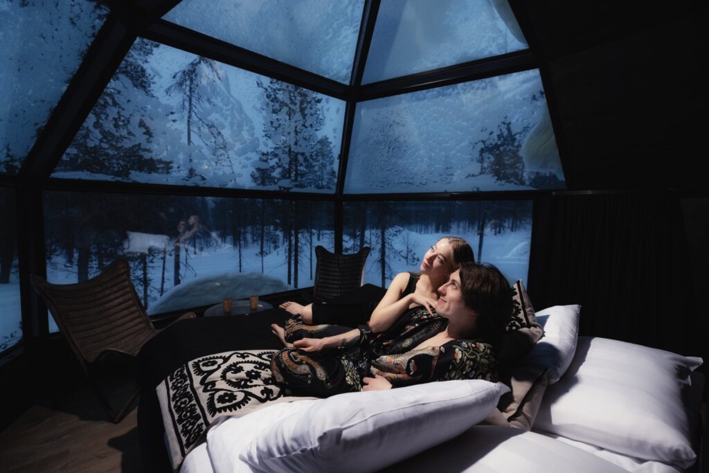 Ein Paar liegt zusammen auf einem Bett in einer glasüberdachten Hütte, lächelt und blickt nach draußen auf eine verschneite Winterlandschaft unter einem dunklen Sternenhimmel. Der gemütliche Raum ist mit gemusterten Decken und modernen Stühlen ausgestattet.