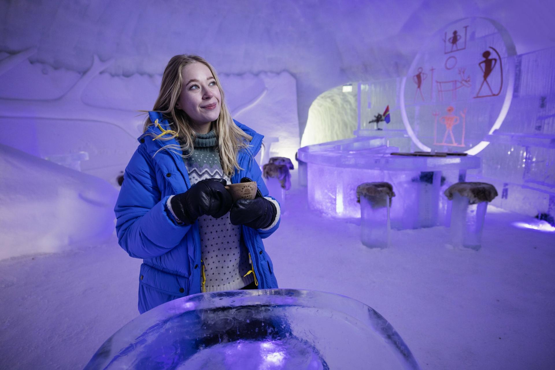 A woman in a blue winter jacket stands inside an ice bar, holding a drink and smiling. The room is made of ice with glowing purple lights and abstract art on the icy walls and furniture.