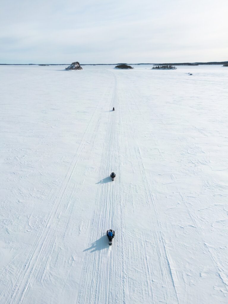 Luftaufnahme von drei Personen, die in einer Reihe auf einem Motorschlitten über einen riesigen, schneebedeckten, zugefrorenen See fahren, auf dem in der Ferne unter einem blassen Himmel kleine Inseln zu sehen sind.