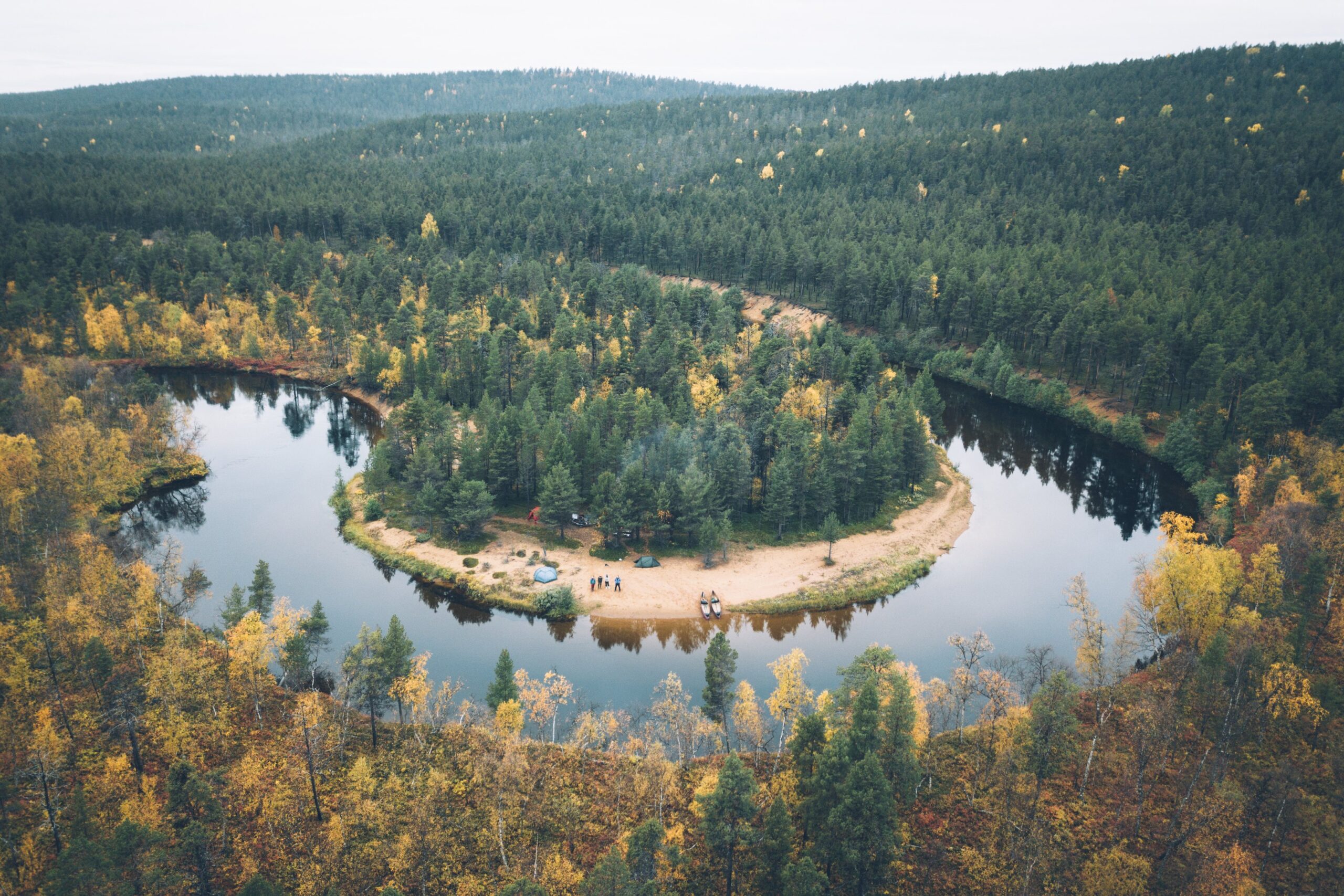 Ilmakuva metsäisestä joen mutkasta ja hiekkasärkästä. Rannalla näkyy ihmisiä ja kanootteja, joita ympäröi tiheä puusto, jossa on alkusyksyn lehdet. Näkymä on rauhallinen ja luonnollinen.