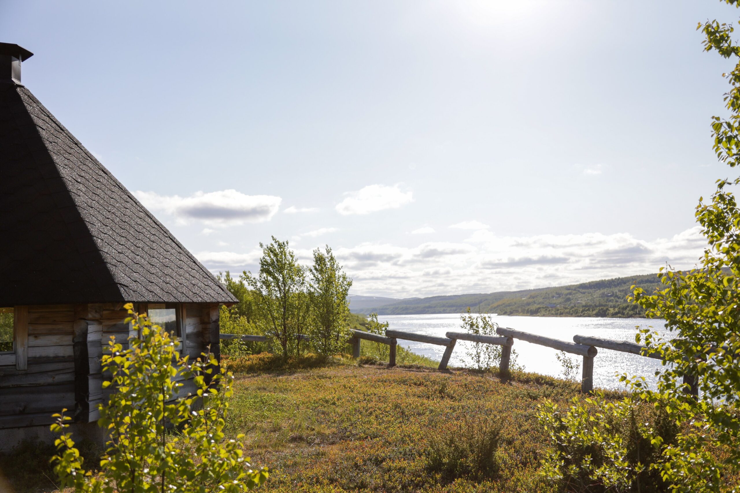 Maalaismainen puinen mökki, jossa on peltikatto, sijaitsee joen rannalla vihreiden pensaiden ja puiden ympäröimänä. Puinen aita kulkee joen rannalla, ja kukkulat levittäytyvät kaukaisuudessa kirkkaan, aurinkoisen taivaan alla.