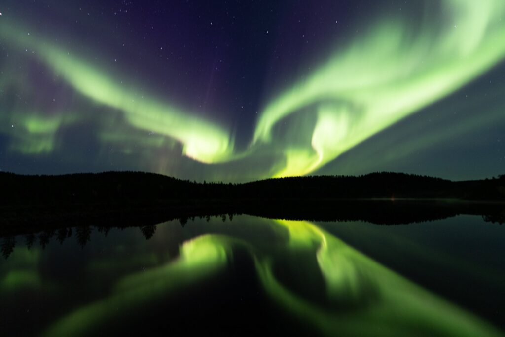 Des aurores boréales vertes et jaunes dansent dans un ciel étoilé, leurs couleurs vibrantes se reflétant dans un lac calme et sombre en contrebas. Une silhouette d'arbres se dessine à l'horizon.