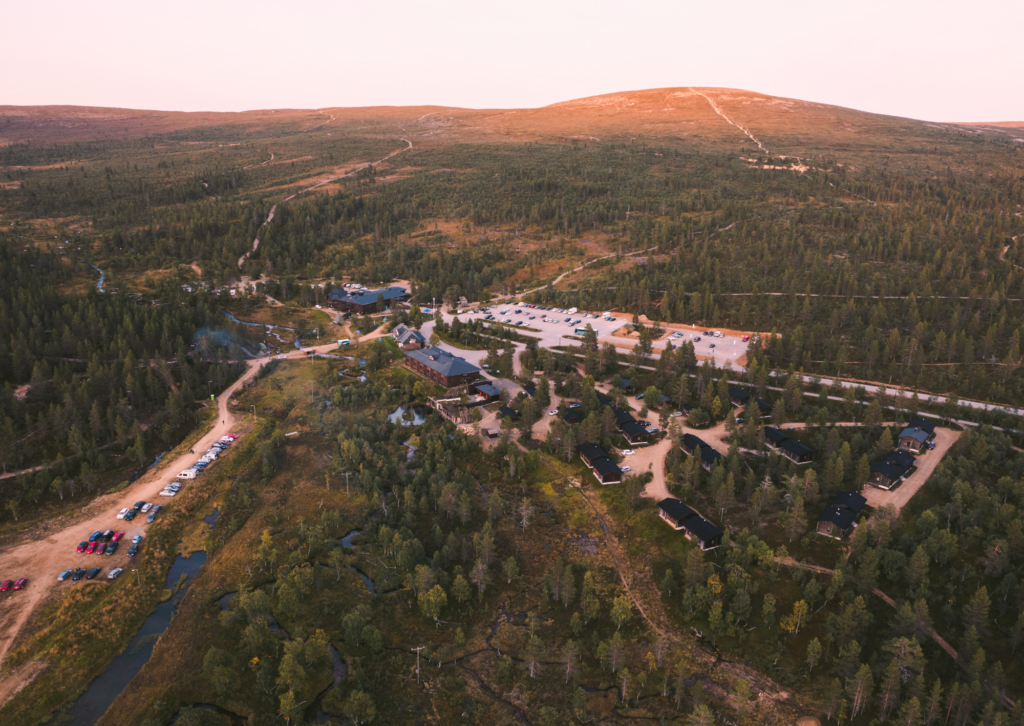 Aerial view of a forested area with scattered buildings, roads, and parked cars, surrounded by rolling hills under a soft pink sky at sunset or sunrise.
