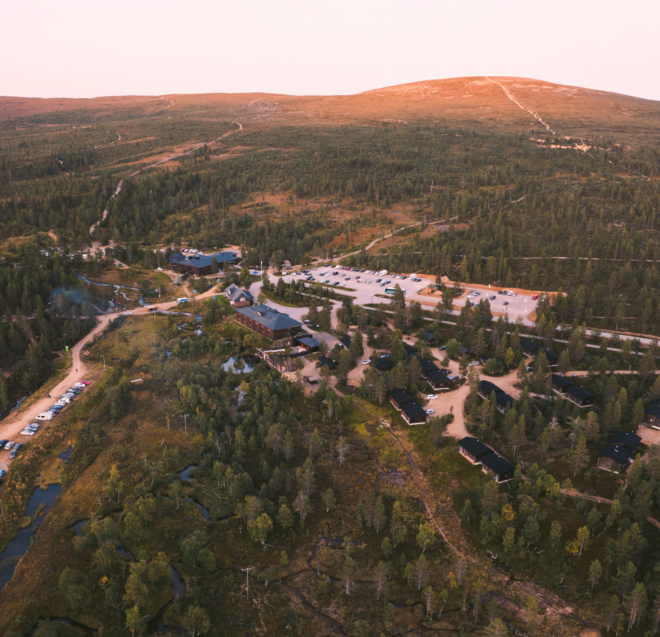 Aerial view of a forested area with scattered buildings, roads, and parked cars, surrounded by rolling hills under a soft pink sky at sunset or sunrise.
