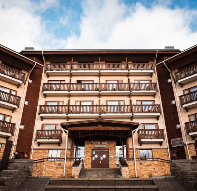 A wide-angle view of a multi-story lodge with wooden balconies, stone accents, and a central entrance under a peaked roof, set against a partly cloudy sky.