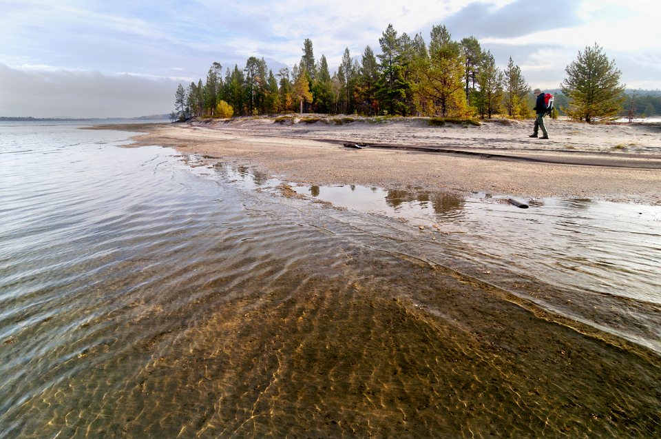 Une personne munie d'un sac à dos rouge marche le long d'un lac sablonneux bordé d'arbres. L'eau est calme et claire, reflétant les arbres et le ciel au-dessus. La scène est paisible et naturelle.