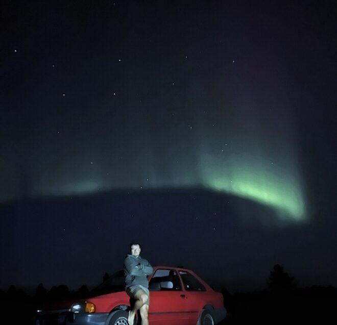 Henkilö istuu punaisen auton konepellillä yöllä ajovalojen valaisemana. Taivaan yllä näkyy elinvoimainen vihreä revontuli, ja taustalla näkyy heikkoja tähtiä.