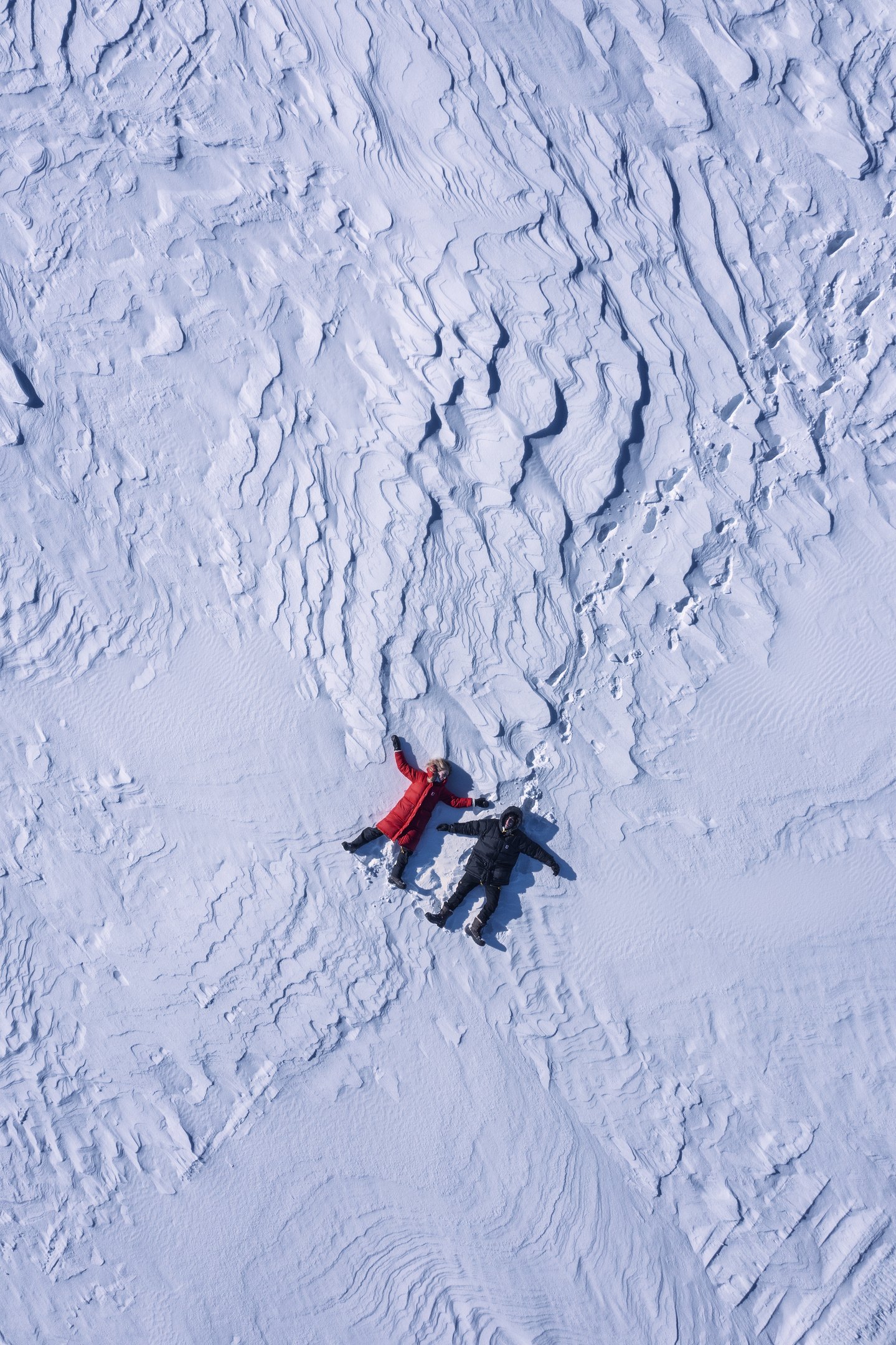 Aerial view of two people lying on snow-covered ground, one in a red coat and the other in dark clothing, surrounded by textured snow patterns and footprints.