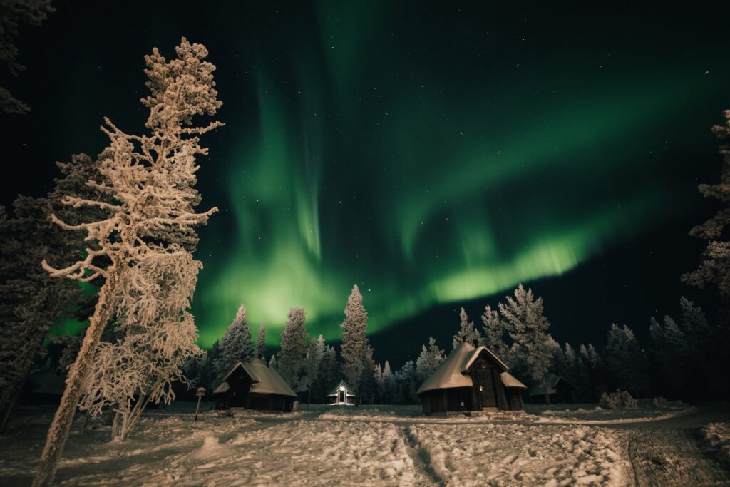 Lumen peittämiä mökkejä ja puita vihreiden revontulien valaiseman yötaivaan alla, pehmeät varjot lumisella maalla ja metsä taustalla.