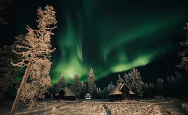 Lumen peittämiä mökkejä ja puita vihreiden revontulien valaiseman yötaivaan alla, pehmeät varjot lumisella maalla ja metsä taustalla.