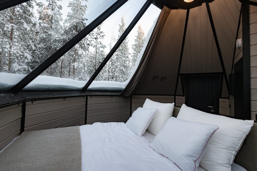 Viihtyisä sänky, jossa on valkoiset vuodevaatteet, on sijoitettu mökin suurten lasi-ikkunoiden alle, joista on selkeä näkymä ulkona olevaan lumiseen metsään. Luonnonvalo täyttää huoneen vinon lasikaton läpi.