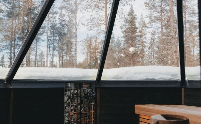 Puurakenteisen saunan sisätiloissa on keskellä metallinen kiuas, ja suurista lasi-ikkunoista avautuu näkymä lumiselle maalle ja korkeille männyille ulkona sinisen taivaan alla.