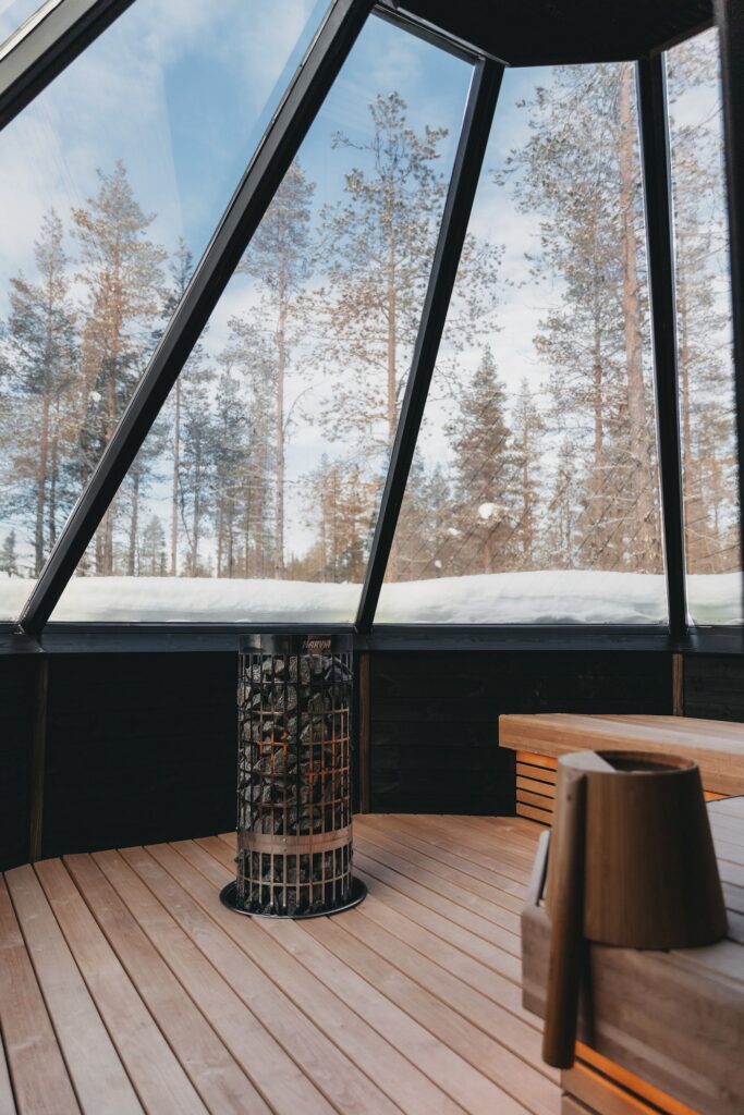 Puurakenteisen saunan sisätiloissa on keskellä metallinen kiuas, ja suurista lasi-ikkunoista avautuu näkymä lumiselle maalle ja korkeille männyille ulkona sinisen taivaan alla.
