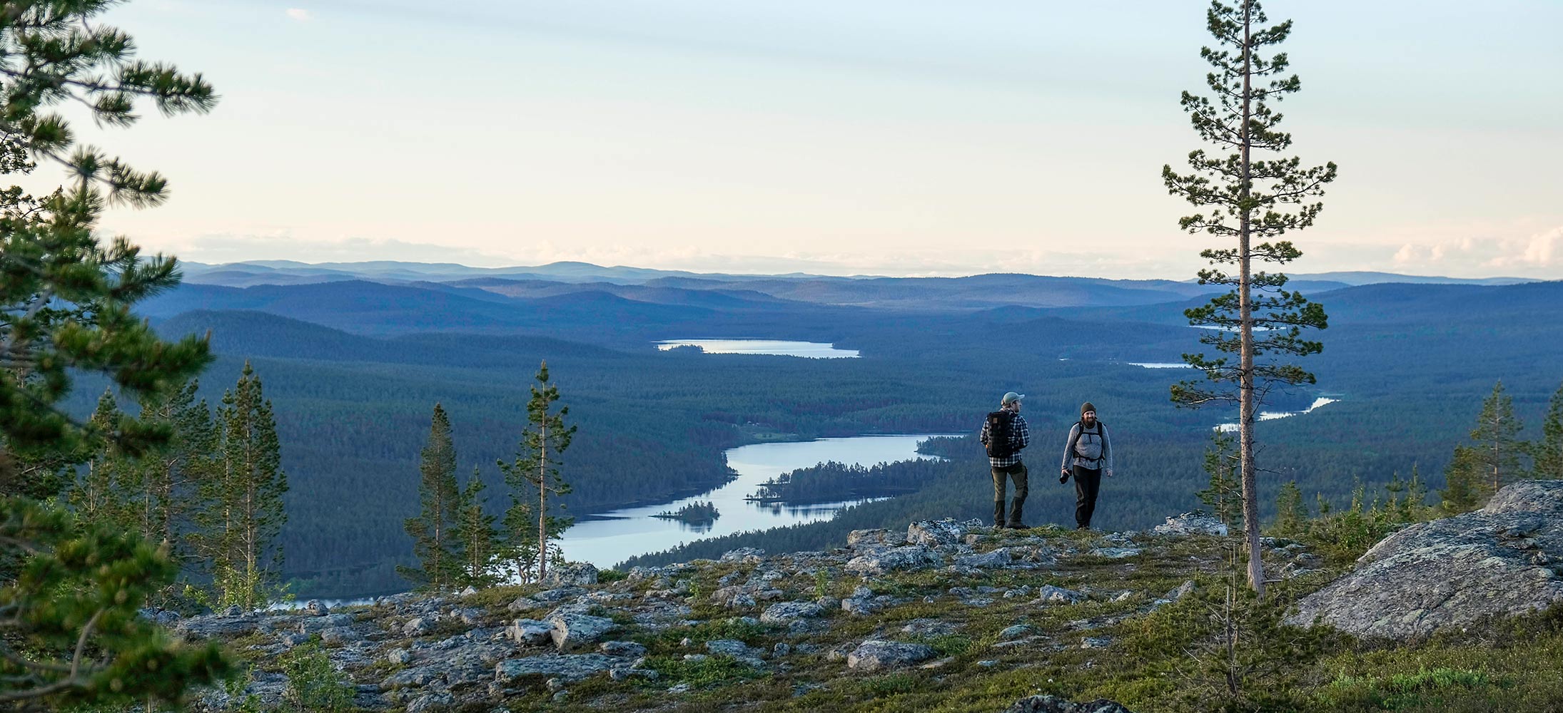 A wide mountain landscape with two hikers walking along a rocky ridge in the foreground. Below them, forest-covered hills stretch into the distance, with winding lakes reflecting the pale evening sky. Tall pine trees frame the scene, and the overall atmosphere is calm and expansive.