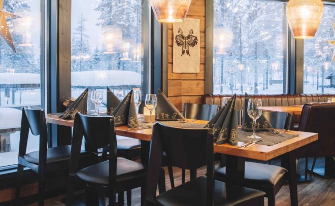 Viihtyisä ravintolan sisustus, jossa on puulattiat ja -katto, mustat tuolit, katettu ruokapöytä, suuret ikkunat, joista näkyy luminen maisema, ja lämmin riippuvalaistus.