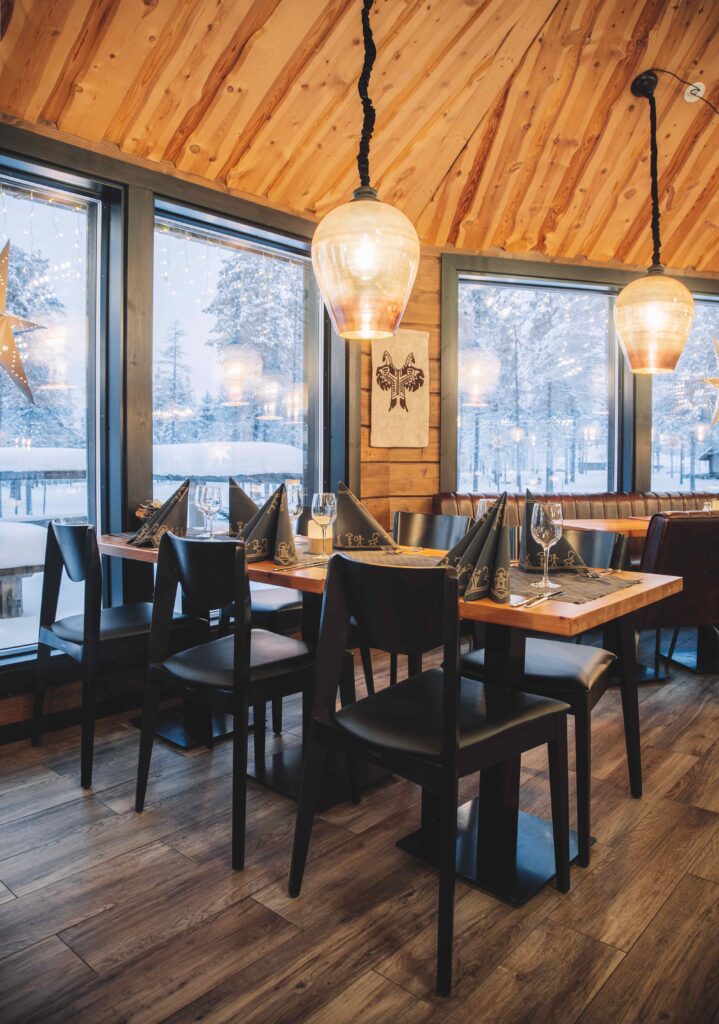 Viihtyisä ravintolan sisustus, jossa on puulattiat ja -katto, mustat tuolit, katettu ruokapöytä, suuret ikkunat, joista näkyy luminen maisema, ja lämmin riippuvalaistus.