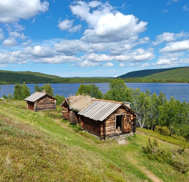 Pienet puiset mökit, joissa on vino katto, istuvat ruohoisella rinteellä, josta on näkymät leveälle, rauhalliselle järvelle, jota ympäröi vihreä metsä sinisen taivaan alla, jossa on hajanaisia valkoisia pilviä.