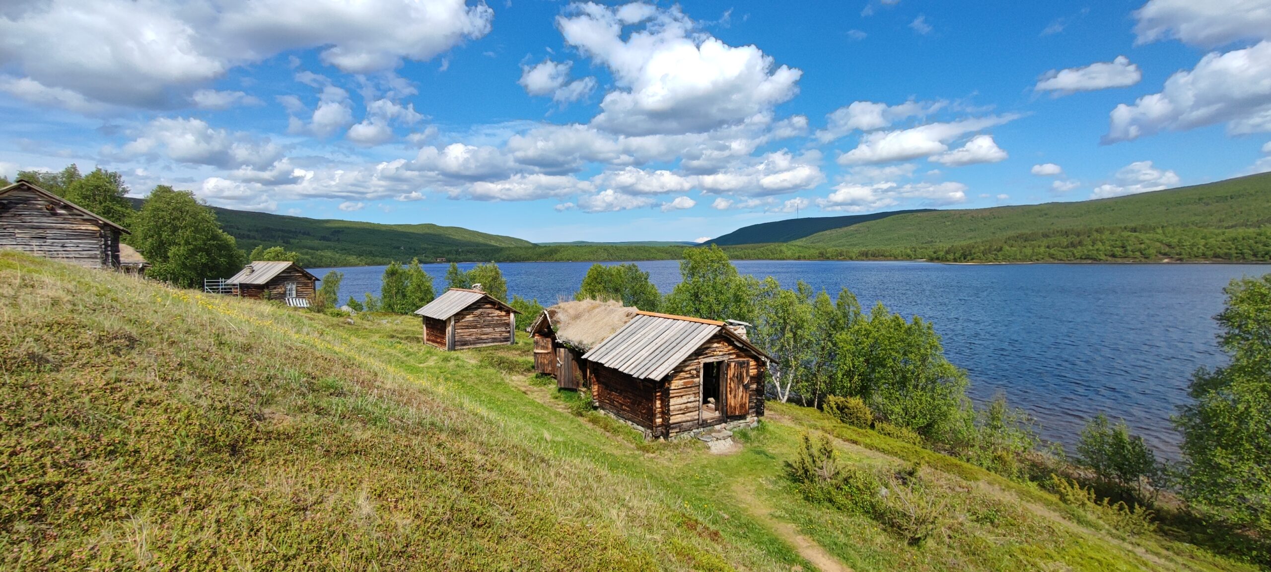 Useita pieniä puisia mökkejä, joissa on ruohokatot, sijaitsee ruohoisella rinteellä, josta avautuu näkymä suurelle järvelle, jota ympäröivät vihreät puut ja kumpuilevat kukkulat kirkkaan sinisen taivaan alla, jossa on hajapilviä.