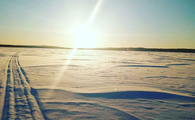 Aurinko paistaa kirkkaasti laajan, lumen peittämän maiseman ylle, jossa on pitkiä varjoja ja kaukaisuuteen johtavia jälkiä kirkkaansinisellä taivaalla.