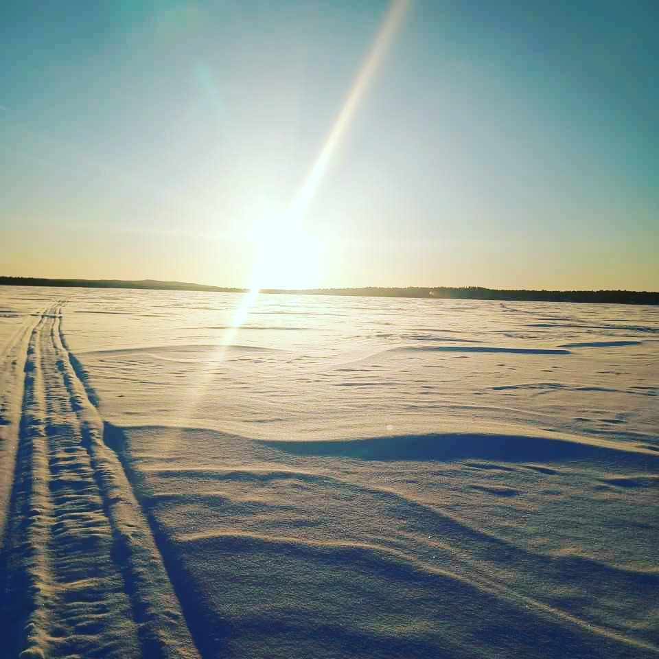Aurinko paistaa kirkkaasti laajan, lumen peittämän maiseman ylle, jossa on pitkiä varjoja ja kaukaisuuteen johtavia jälkiä kirkkaansinisellä taivaalla.