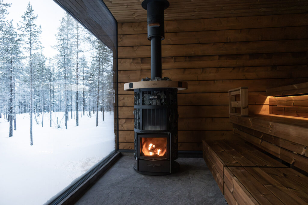Viihtyisä puusauna, jossa on polttava kiuas, puupenkit ja suuri ikkuna, josta avautuu luminen metsänäkymä ulos. Lämmin sisävalo muodostaa kontrastin kylmään, talviseen maisemaan.