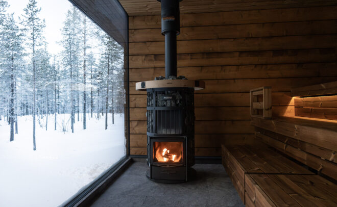 Viihtyisä puusauna, jossa on polttava kiuas, puupenkit ja suuri ikkuna, josta avautuu luminen metsänäkymä ulos. Lämmin sisävalo muodostaa kontrastin kylmään, talviseen maisemaan.