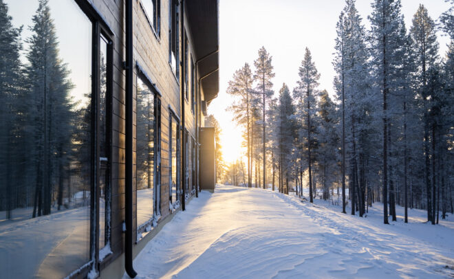 Moderni rakennus, jossa on suuret ikkunat, seisoo lumisessa maisemassa, jossa on korkeita mäntyjä, kun aurinko nousee tai laskee, heittää lämpimän kultaisen valon lumen päälle ja luo pitkiä varjoja.