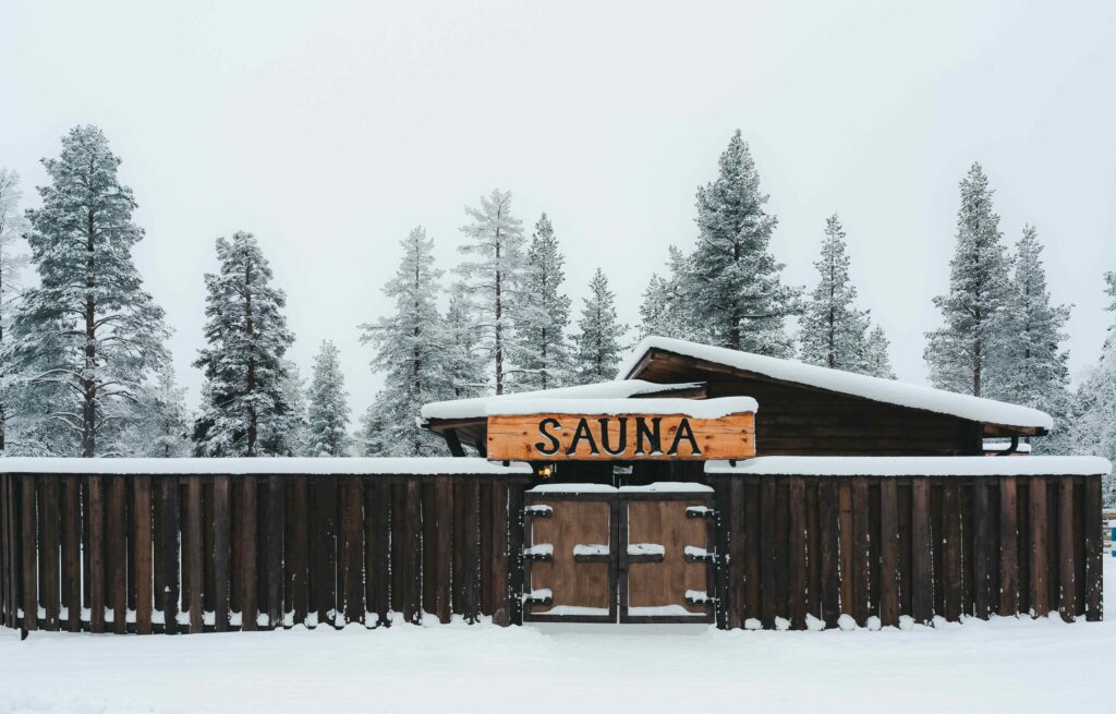Puinen saunarakennus, jota ympäröi lumipeitteinen aita ja puita, portin yläpuolella suuri SAUNA-kyltti talvimaisemassa.