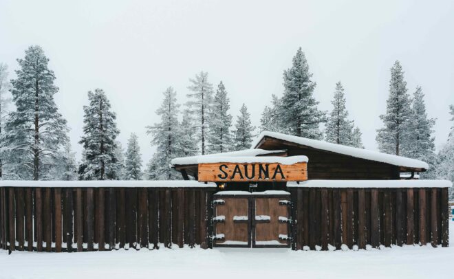 Puinen saunarakennus, jota ympäröi lumipeitteinen aita ja puita, portin yläpuolella suuri SAUNA-kyltti talvimaisemassa.