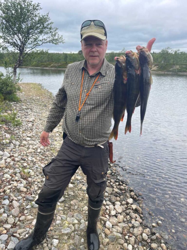 Ulkoiluvaatteisiin pukeutunut mies seisoo kivikkoisella järvenrannalla ja pitelee toisessa kädessään neljää kalaa. Hänellä on saappaat, ruudullinen paita ja lippis, ja taustalla on pilvinen taivas ja puita.