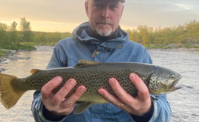Siniseen takkiin ja lippikseen pukeutunut henkilö pitelee molemmilla käsillään suurta täplikäs kalaa joen rannalla, taustalla puita ja pilvinen taivas.