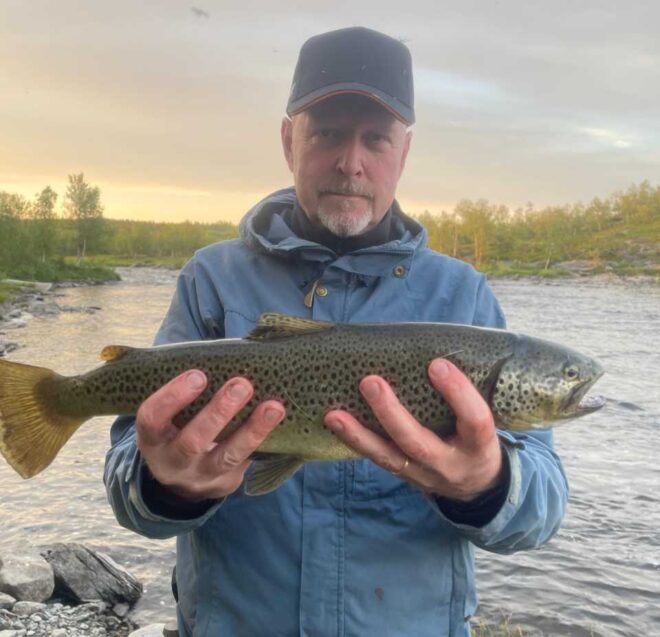 Siniseen takkiin ja lippikseen pukeutunut henkilö pitelee molemmilla käsillään suurta täplikäs kalaa joen rannalla, taustalla puita ja pilvinen taivas.