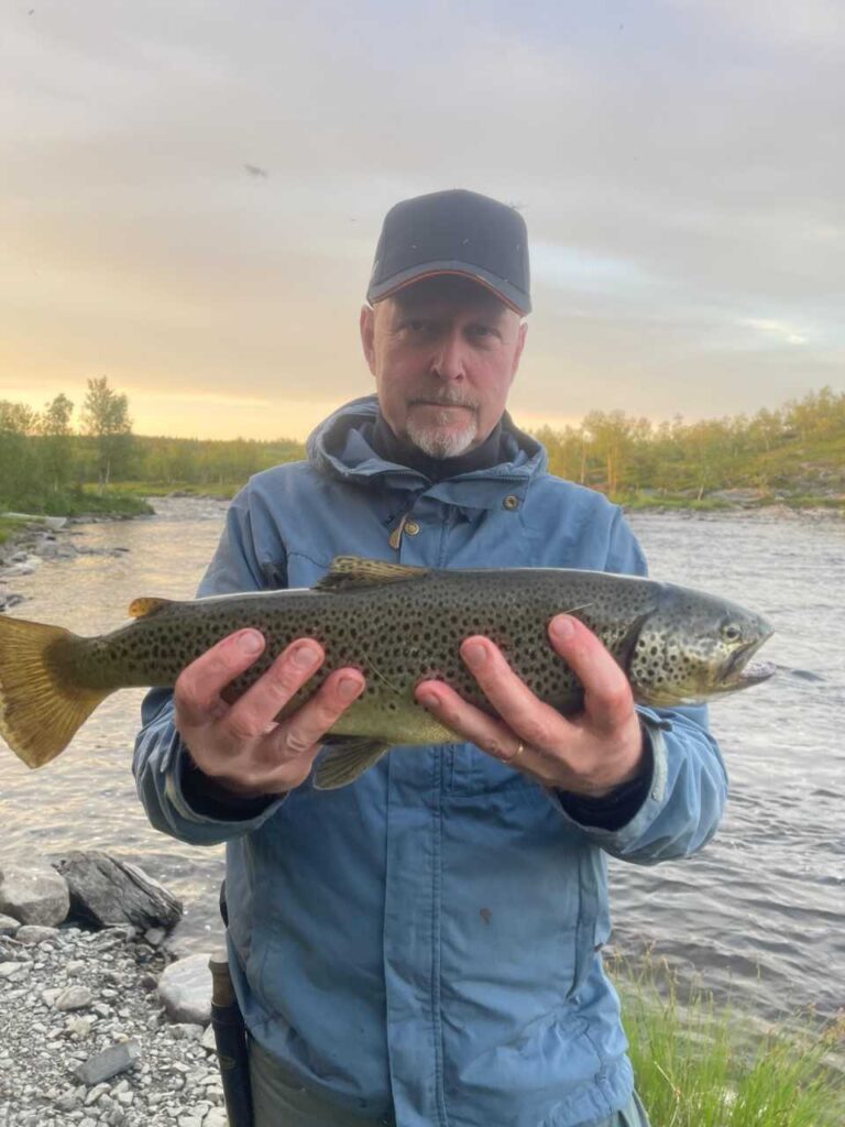 Siniseen takkiin ja lippikseen pukeutunut henkilö pitelee molemmilla käsillään suurta täplikäs kalaa joen rannalla, taustalla puita ja pilvinen taivas.